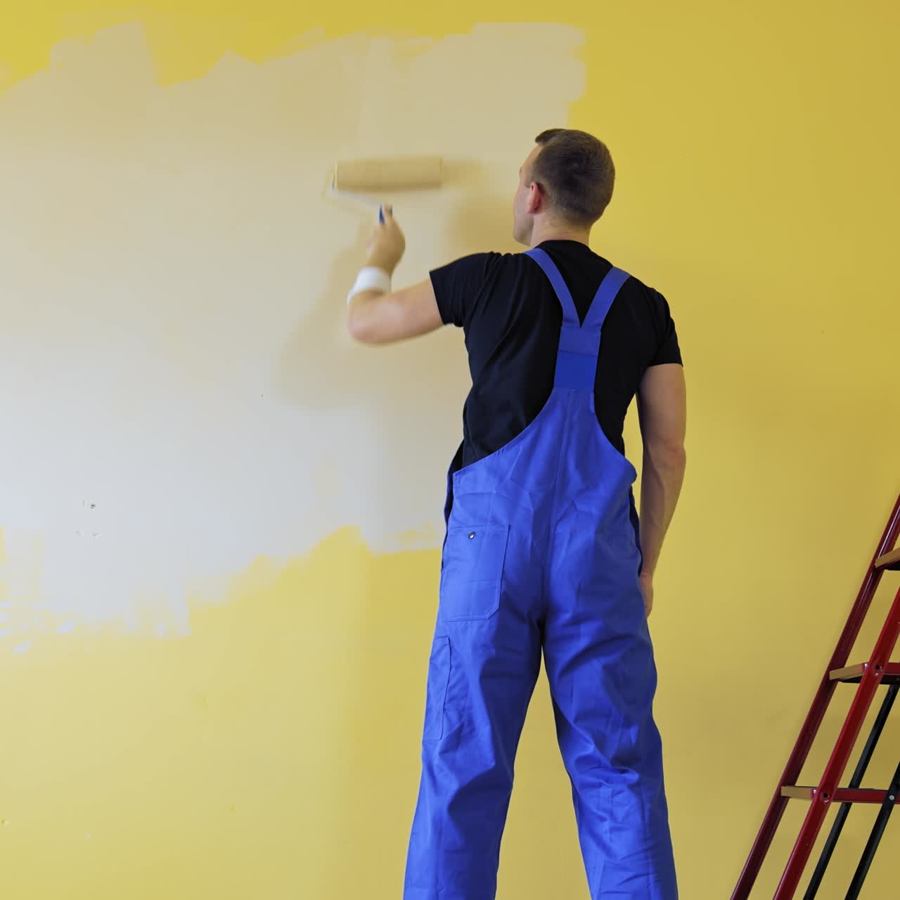 Painter doing the makeover indoors. Male worker painting wall with a paint roller next to the ladder. Improvement of a room interior. Back view