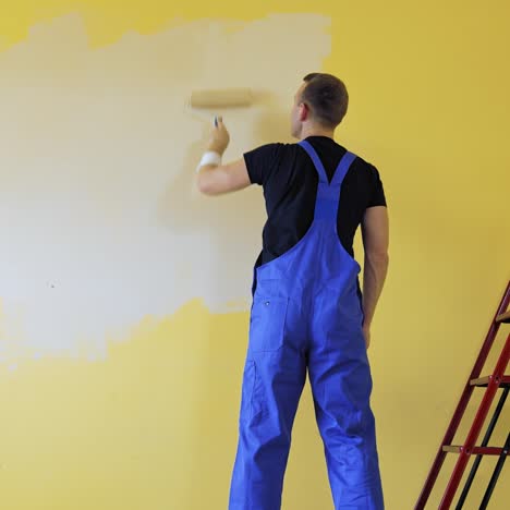 painter doing the makeover indoors. male worker painting wall with a paint roller next to the ladder. improvement of a room interior. back view