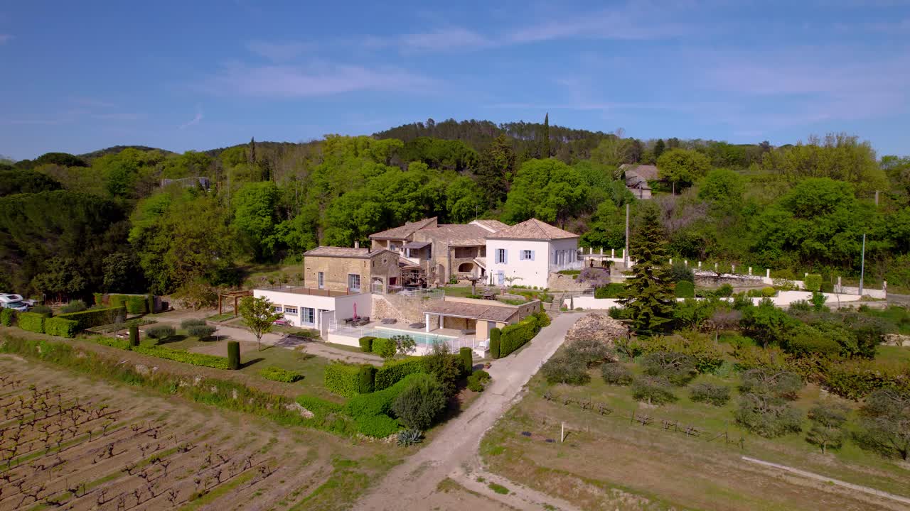 drone aerial shot of a beautiful property in the south of france with several outbuilding, light roofs and wall, a swimming pool and vegetation all around, with the fields in front in the foreground,