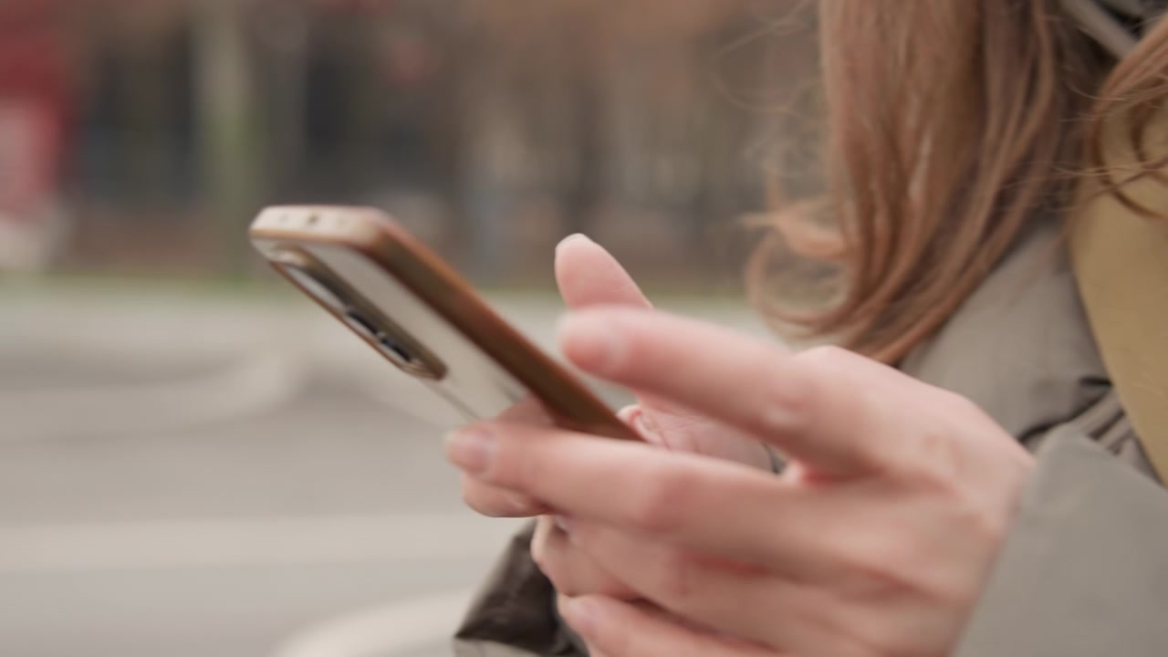 Primo piano di mani che tengono un telefono all'aperto, pendolare urbano concentrato sulla messaggistica e sullo scorrimento delle notifiche mentre aspetta per strada con veicoli di passaggio, cappotto neutro e custodia del telefono visibili, morbido