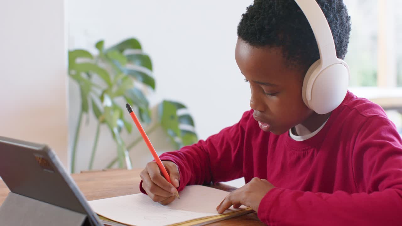African American boy tapping tablet for school prompts and writing in notebook at home dining table