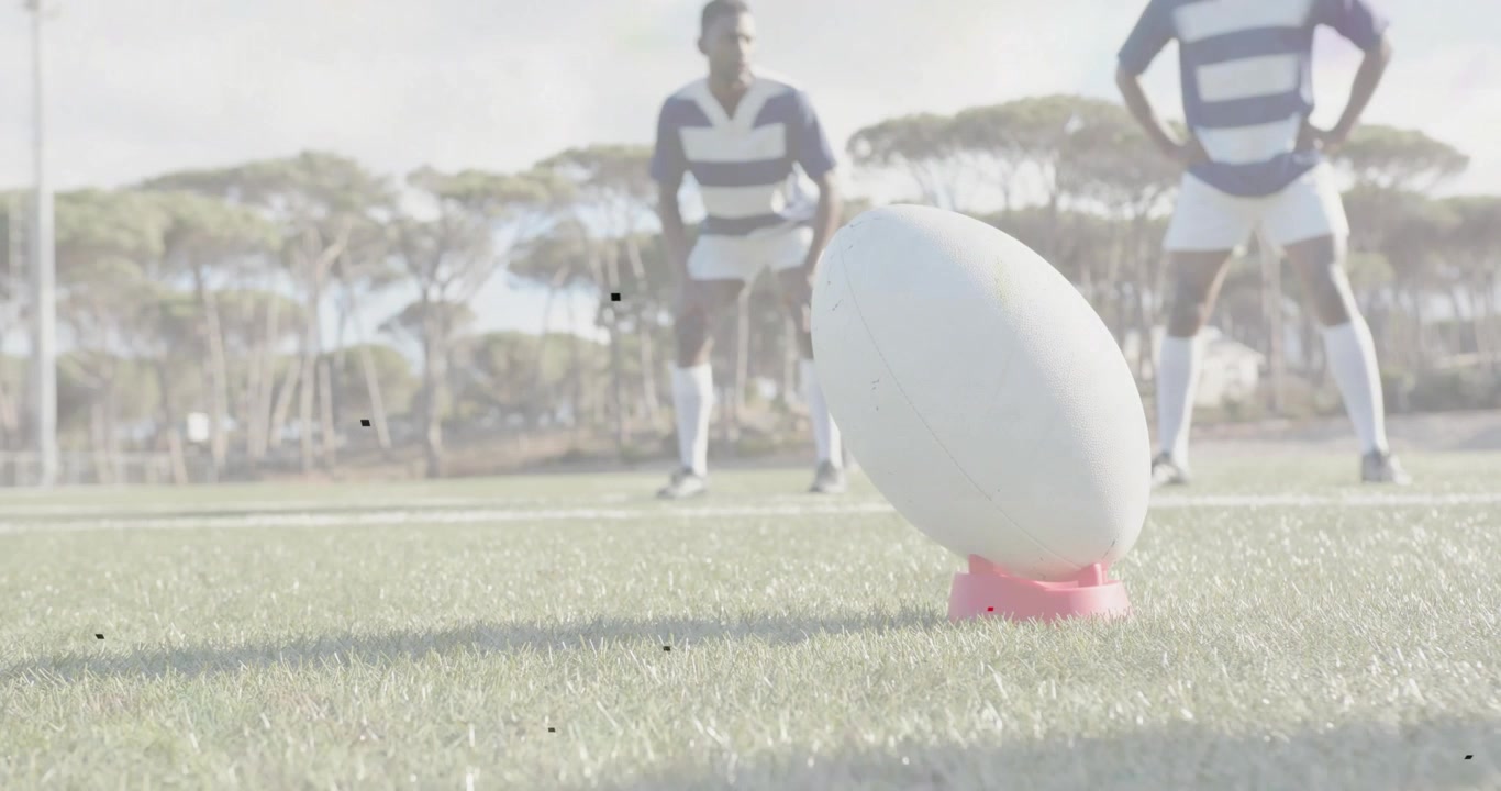 Dwóch mężczyzn obserwuje piłkę do rugby opartą na różowej podstawce, przygotowując się do kopnięcia z miejsca, podczas gdy mijający obiekt rzuca cień.