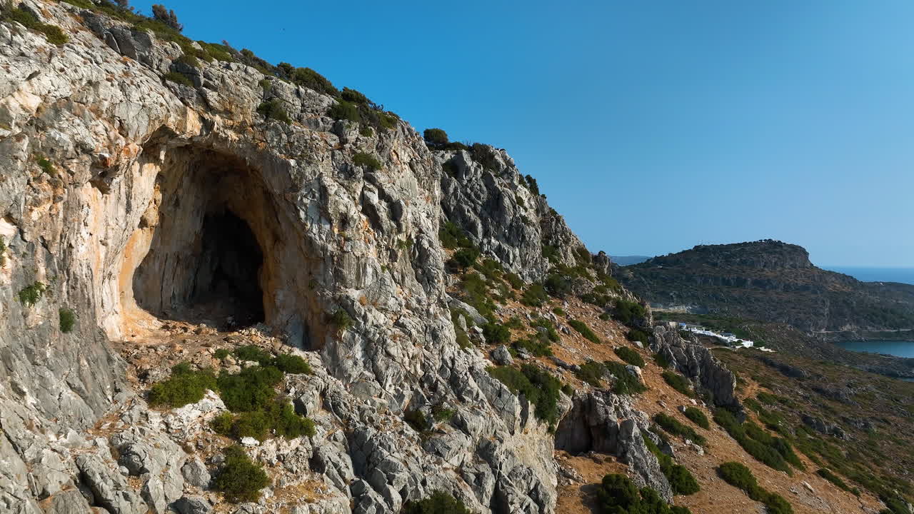 Drone passing a cave with hikers inside, rocky coast of Rhodes, sunny Greece
