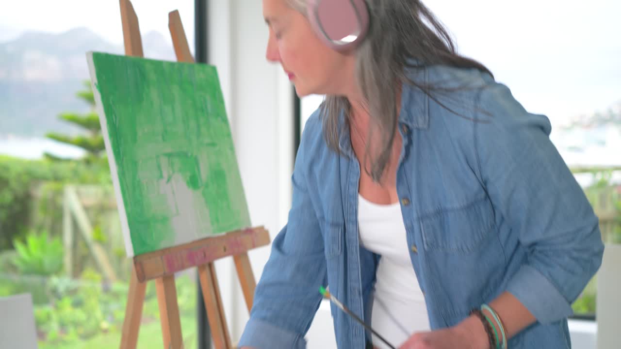 En ældre kvinde inspicerer lærredet og maler derefter grønne strøg som grundfarve ved staffeliet med en palet.