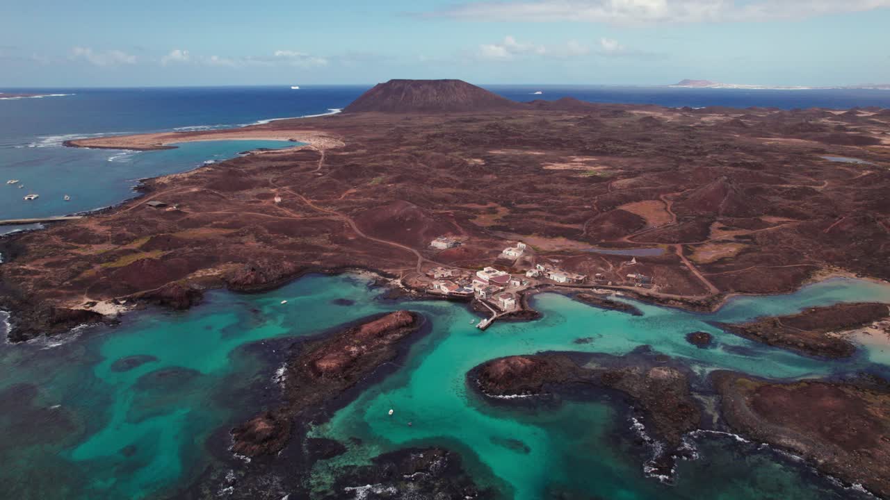 4K aerial drone view of Puertito Isla de Lobos, Fuerteventura, featuring turquoise waters, rocky coastlines, volcanic terrain, and distant mountains that showcase coastal beauty and serene landscapes