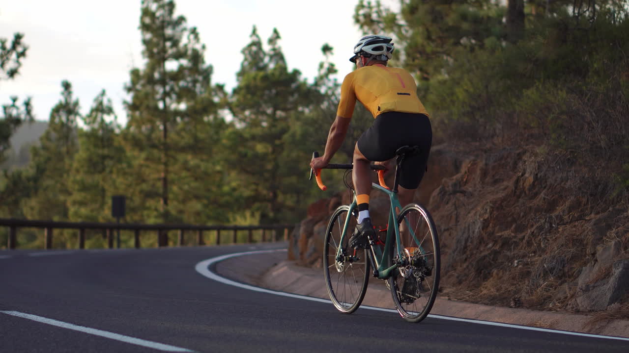 A male individual, dressed in a yellow t-shirt, is cycling along a road situated at a high altitude in the mountains using a sports road bike