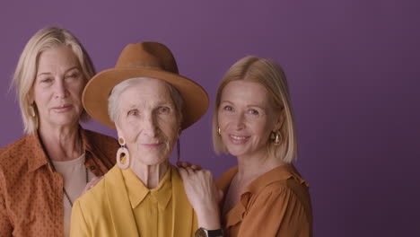 senior woman in hat and mustard shirt poses with two blonde mature women on purple background