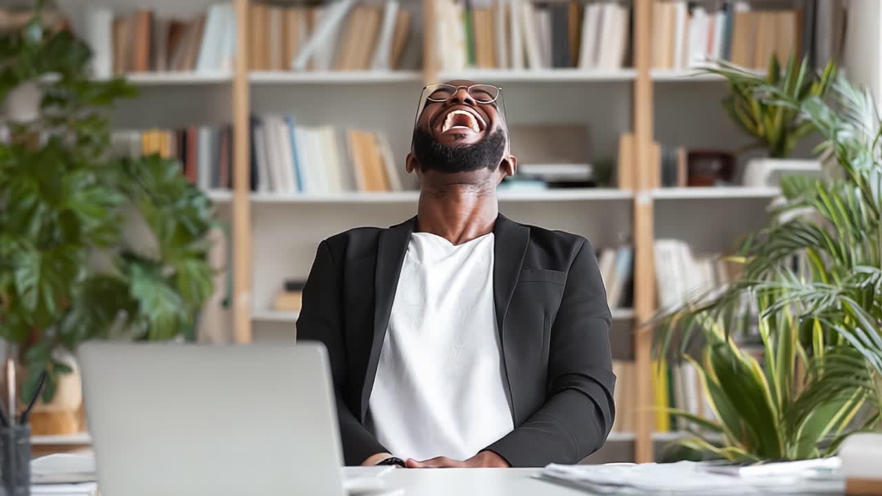 Un uomo gioioso che si gode un momento spensierato alla sua scrivania, mostrando una risata genuina mentre è circondato da uno spazio di lavoro vivace e organizzato, ricco di verde.