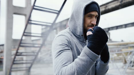 close-up view of handsome bearded caucasian man in grey hoodie and boxing to the camera outdoors an abandoned factory on a cloudy morning