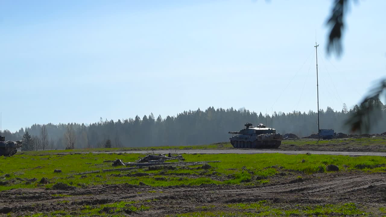 Rare video of multiple British Challenger 2 4034 tanks aiming at enemy troops on a green grasy field while sun is shining during summer. Filmed from behind some bokeh blurry forest branches moving.
