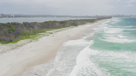 Drone-Aéreo-Sobrevuela-La-Hermosa-Playa-Con-La-Ciudad-En-Segundo-Plano