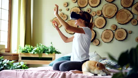 smiling mixed race girl is using augmented reality glasses moving hands sitting on bed in modern apartment with adorable dog beside her. technology and fun concept.