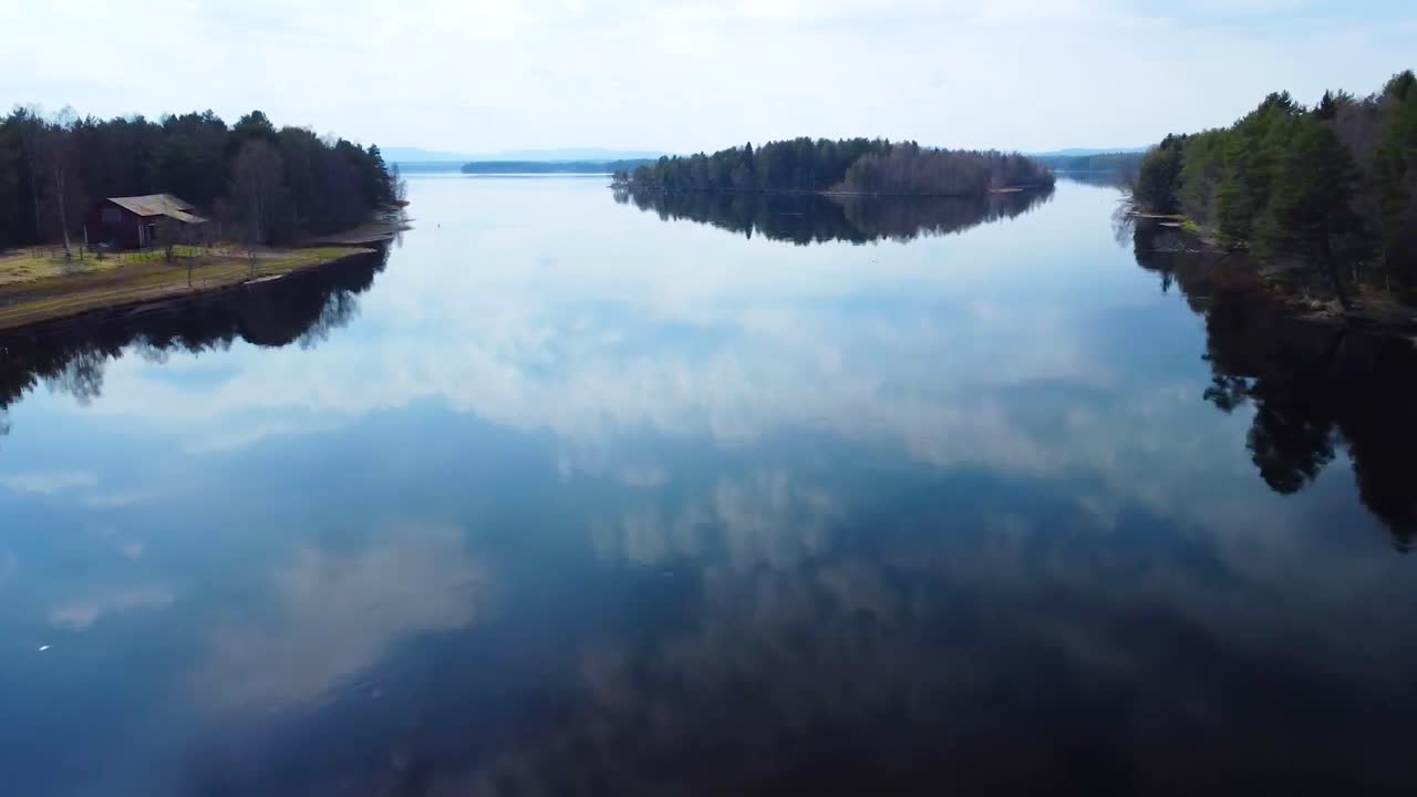 Aerial low flyover over a reflective and glassy blue lake in Sweden during dusk or dawn while the water is reflecting white fluffy clouds and grassy shoreline that have tall pine trees and a cabin.
