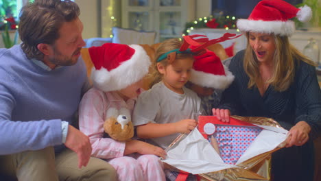 family wearing santa hats sitting on sofa at home opening christmas gifts