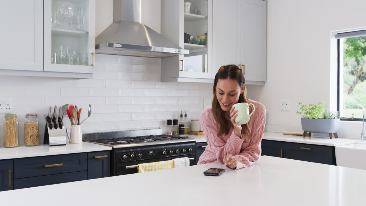 Smiling woman in kitchen enjoying coffee while checking smartphone messages, copy space