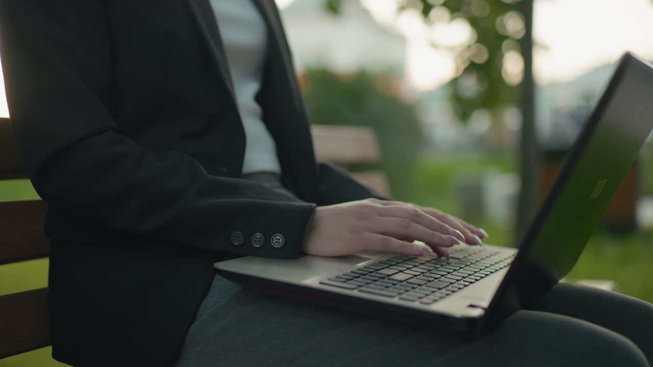Close up partial view of woman in official wear typing on laptop with polished nails seated outdoors on bench in natural environment with blur background featuring greenery and unclear structures