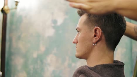 vista lateral de un joven hombre caucásico guapo con perforación en la oreja recibiendo su cabello vestido y peinado por un barbero barbudo en un
