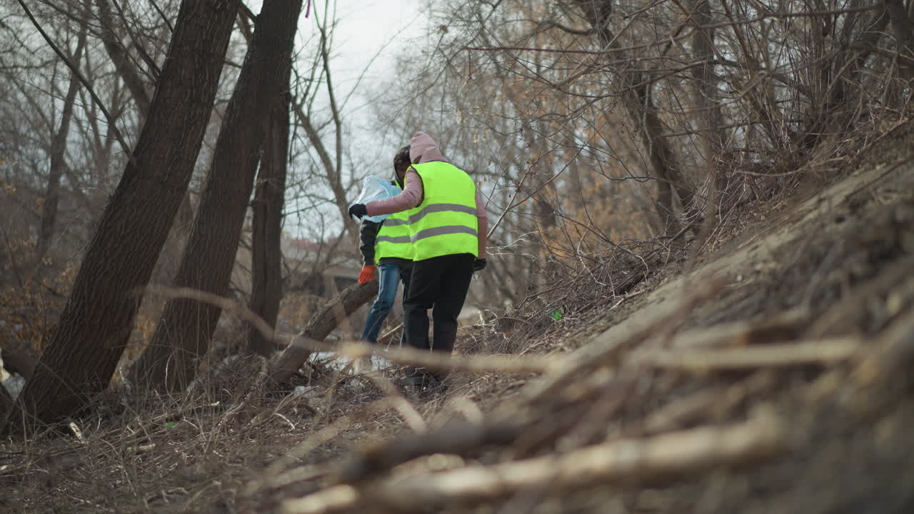 Two volunteers in reflective safety vests and gloves removing large debris and trash from riverbank during outdoor cleanup, placing waste into blue bags to protect nature, support environmental conservation