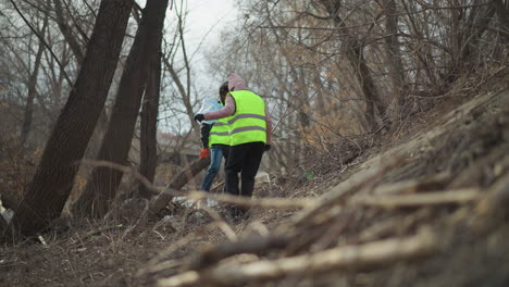 two volunteers in reflective safety vests and gloves removing large debris and trash from riverbank during outdoor cleanup, placing waste into blue bags to protect nature, support environmental conservation