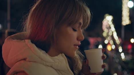 lady sipping hot beverage at night park surrounded by christmas lights closeup.