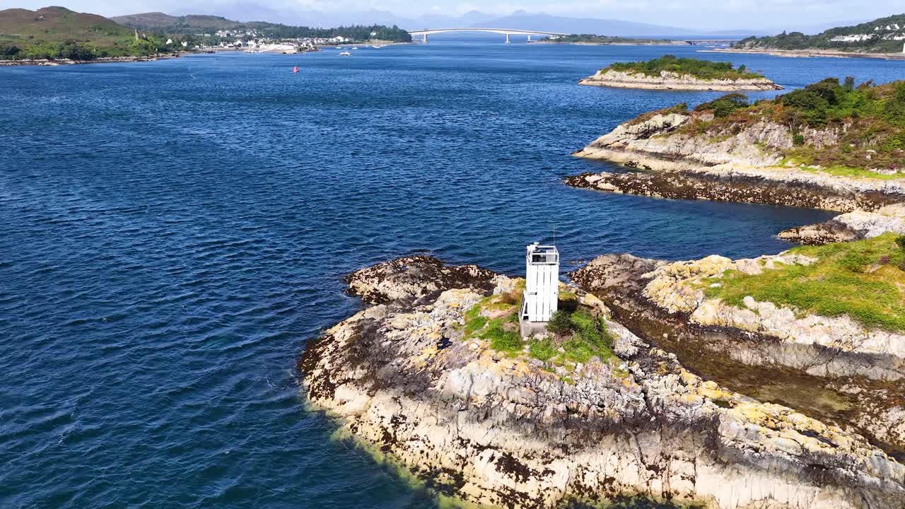 Drone glides over rocky island with lighthouse, blue sea, and distant Scottish mainland under sunlight