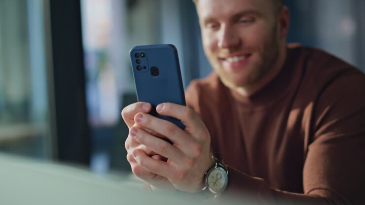 Primo piano di un uomo sorridente che tiene in mano uno smartphone nel suo ufficio domestico. Imprenditore che usa il telefono