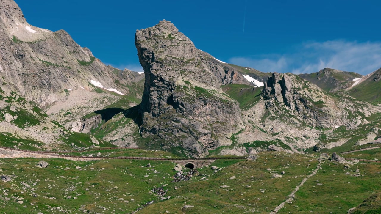 Steinfylt fjellandskap med grønne skråninger og tunnel under klar blå himmel, en sommerdag.