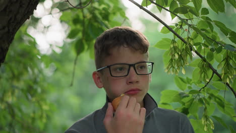joven con una camiseta gris y gafas comiendo un bocadillo tranquilamente bajo un árbol, aparece profundamente en pensamiento, rodeado de hojas verdes exuberantes en un ambiente al aire libre tranquilo