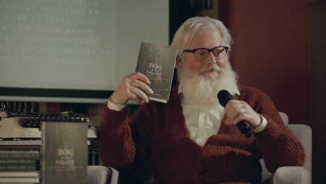 senior writer holding book and talking in mic from stage at book launch