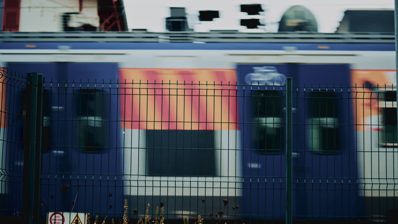 Blurred high speed train with colorful graphics rushes past behind a metal fence, conveying motion, travel and urban transportation infrastructure