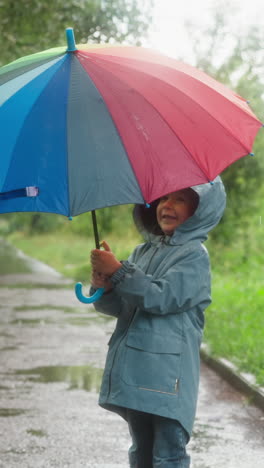 happy child opens umbrella in rainy park. smiling little boy in hooded coat holds colorful parasol walking in spring garden. bright accessory for gloomy weather