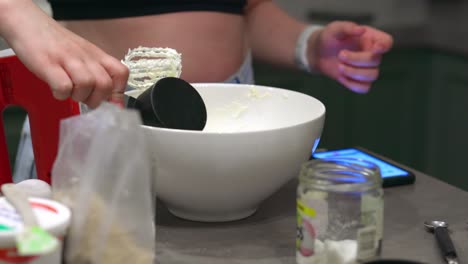 una mujer usa una taza medidora negra para verter azúcar en un tazón blanco, luego procede a recoger una batidora de mano roja para mezclar los ingredientes para hornear y hacer un pastel de queso