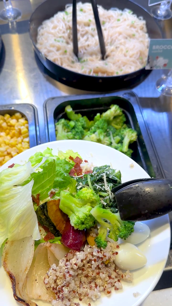 Ovenfra ses en tang, der placerer grøn broccoli på en tallerken ved en farverig salatbar.