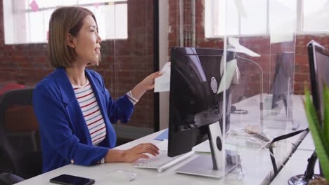 Asian-businesswoman-sitting-at-desk-using-computer-and-passing-document-over-sneeze-guard