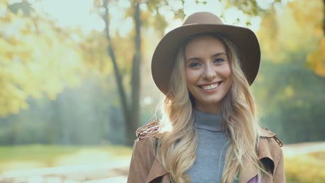 Close-up-view-of-young-blonde-woman-wearing-a-hat-and-smiling-at-the-camera-in-the-park-in-the-morning