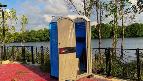portable toilet with open door placed near river walkway—great for editorial, public service, construction, or environmental themes