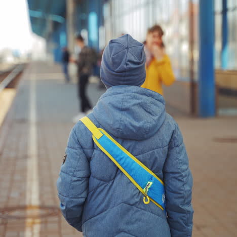 back side view of a boy standing alone on a platform and waiting for the train. little boy in a gray cap and a jacket with a backpack on the railway station.