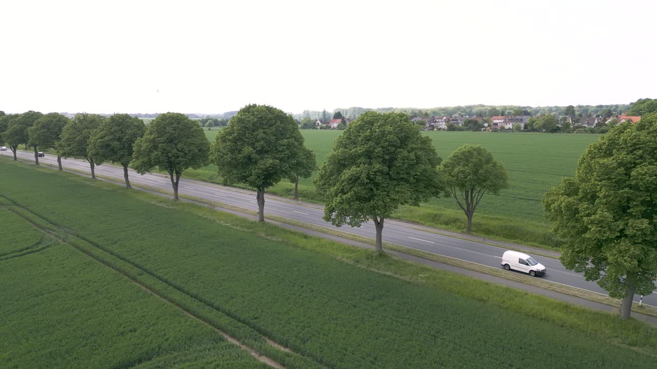 Aerial drone shot of a road with cars and bus passing by, surrounded by green farmlands along rural countryside of Brunswick, Germany on a cloudy day