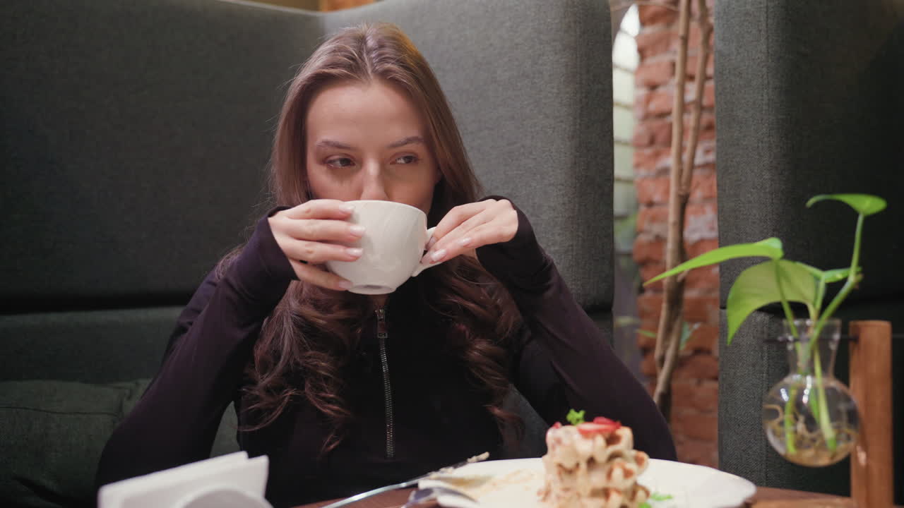 Lady holds white ceramic cup with both hands while sipping latte in stylish booth featuring decorative vase, lush green plant, and partially eaten waffles, evoking calm and cozy indoor coffee moment