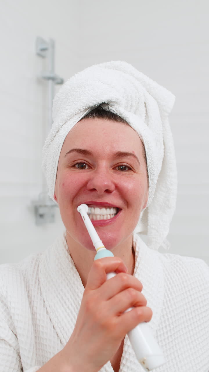 Middleaged woman smiling brushing teeth carefully using electric toothbrush at bathroom mirror