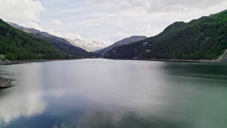 aerial flying over lai da marmorera reservoir surrounded by forest trees with snow capped mountains seen in distance background