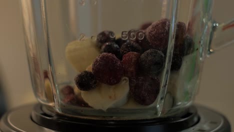 woman puts a pieces of berries and banana in the bowl of a blender, close up shot