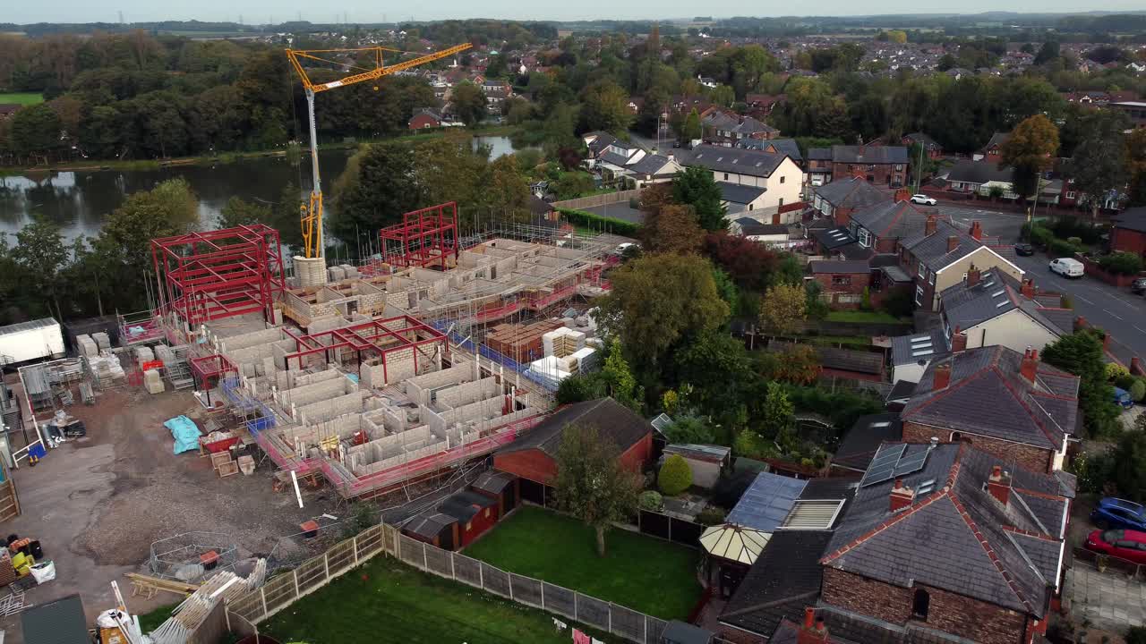 Aerial view crane building waterside care home construction framework in rural British village next to fishing lake, pull away shot
