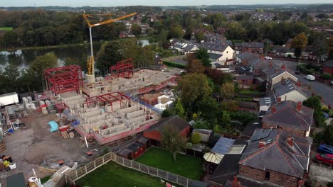 Aerial-view-crane-building-waterside-care-home-construction-framework-in-rural-British-village-next-to-fishing-lake,-pull-away-shot