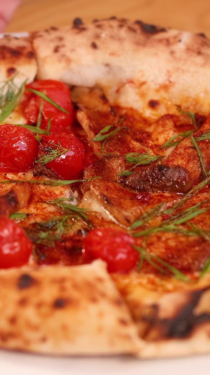 Primo piano di mani che servono una pizza cotta nel forno a legna con pomodorini e erbe fresche.