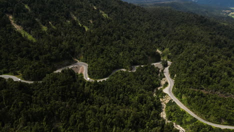 Toma-Aérea-De-Una-Carretera-Con-Curvas-Entre-Un-Denso-Bosque-Montañoso-Del-Oeste-De-Nueva-Zelanda