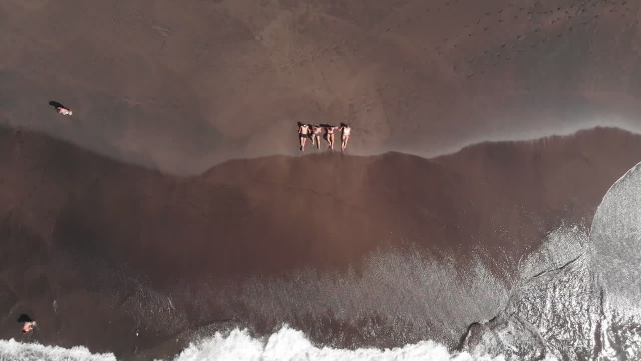 People laying on black sand beach relaxing as the waves roll in - birds eye view descending, Gui Gui Beach, las Palmas de Gran Canaria