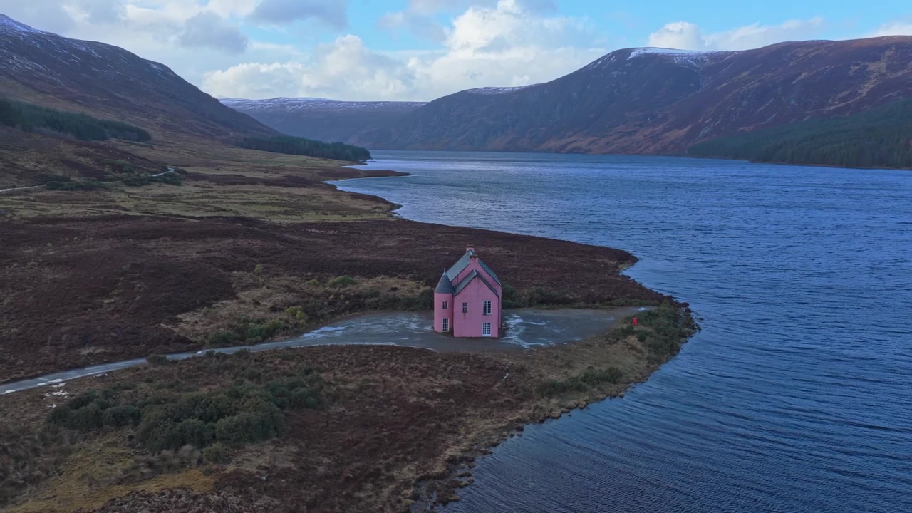 Drohnenaufnahme, die rückwärts fliegt und die atemberaubende Landschaft um ein abgelegenes rosa Haus am Ufer des Loch Glass in Schottland enthüllt