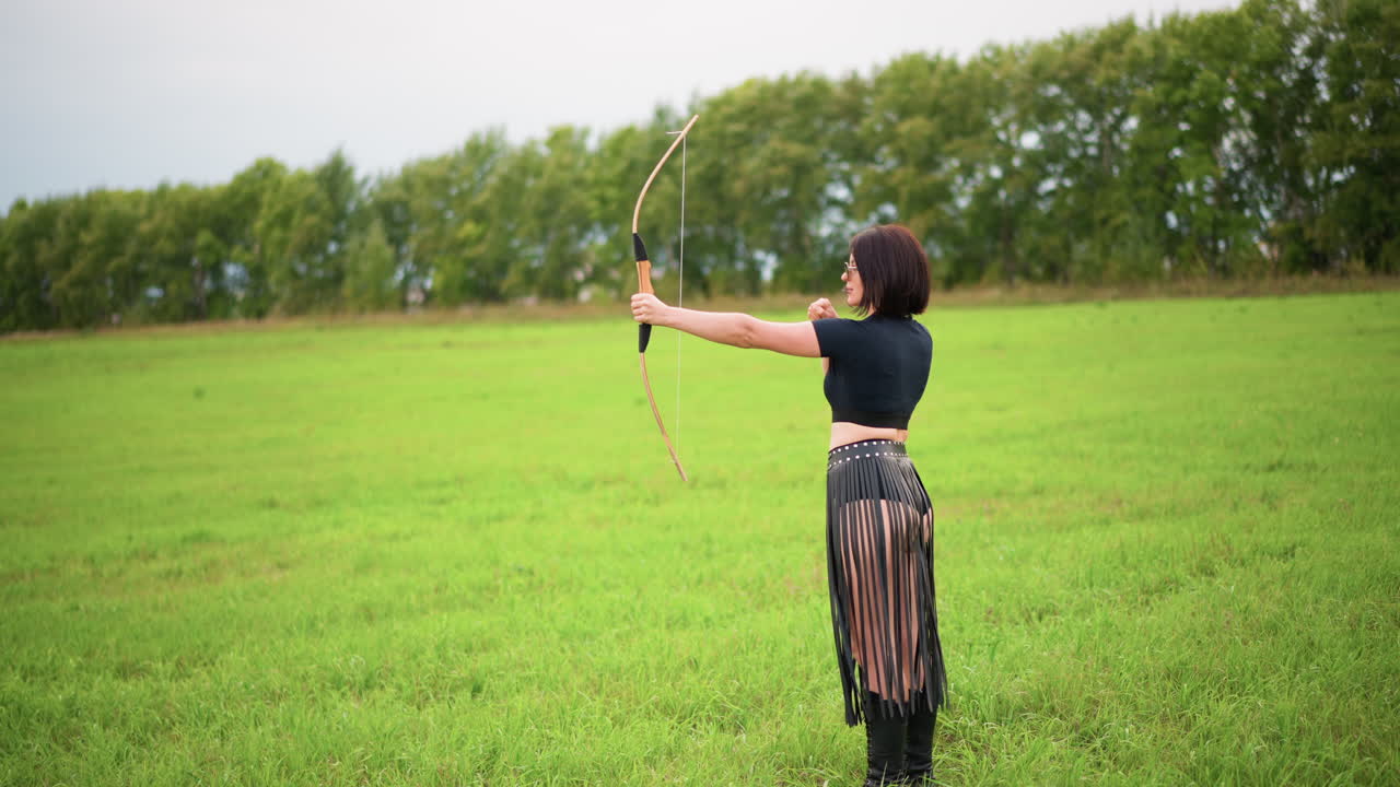 female archer taking aim in meadow with full draw and poised release, leather fringe skirt and boots, side profile silhouette against treeline, steady posture and cinematic motion, athletic