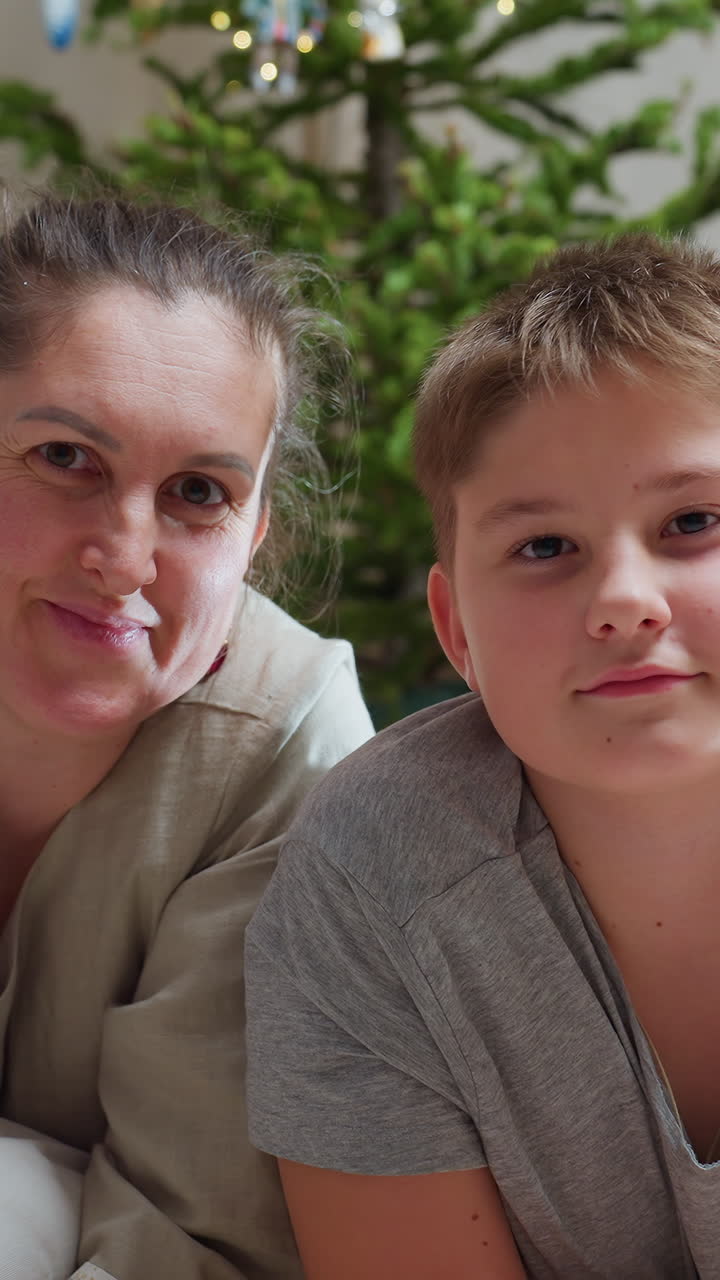 Madre blanca abrazando a su hijo junto a un árbol, con luz suave matinal, luces de hadas con efecto bokeh, cojines de suelo y adornos festivos; nariz y frente se tocan, con sonrisas dulces y un primer plano espontáneo que capta la calidez.